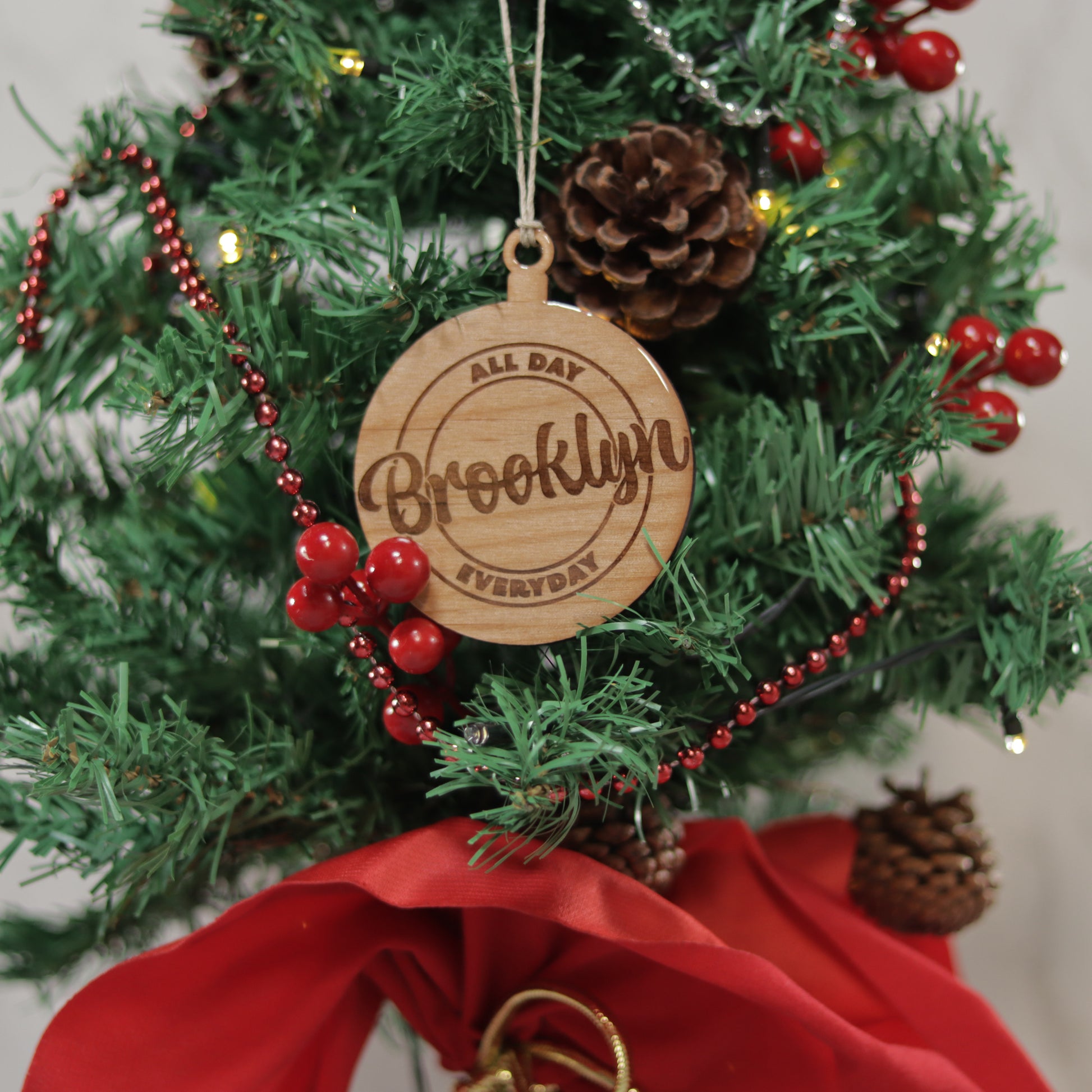 Round wood ornament that is coated with UV resin on a mini Christmas tree.The words Brooklyn all day everyday is etched in the ornament.